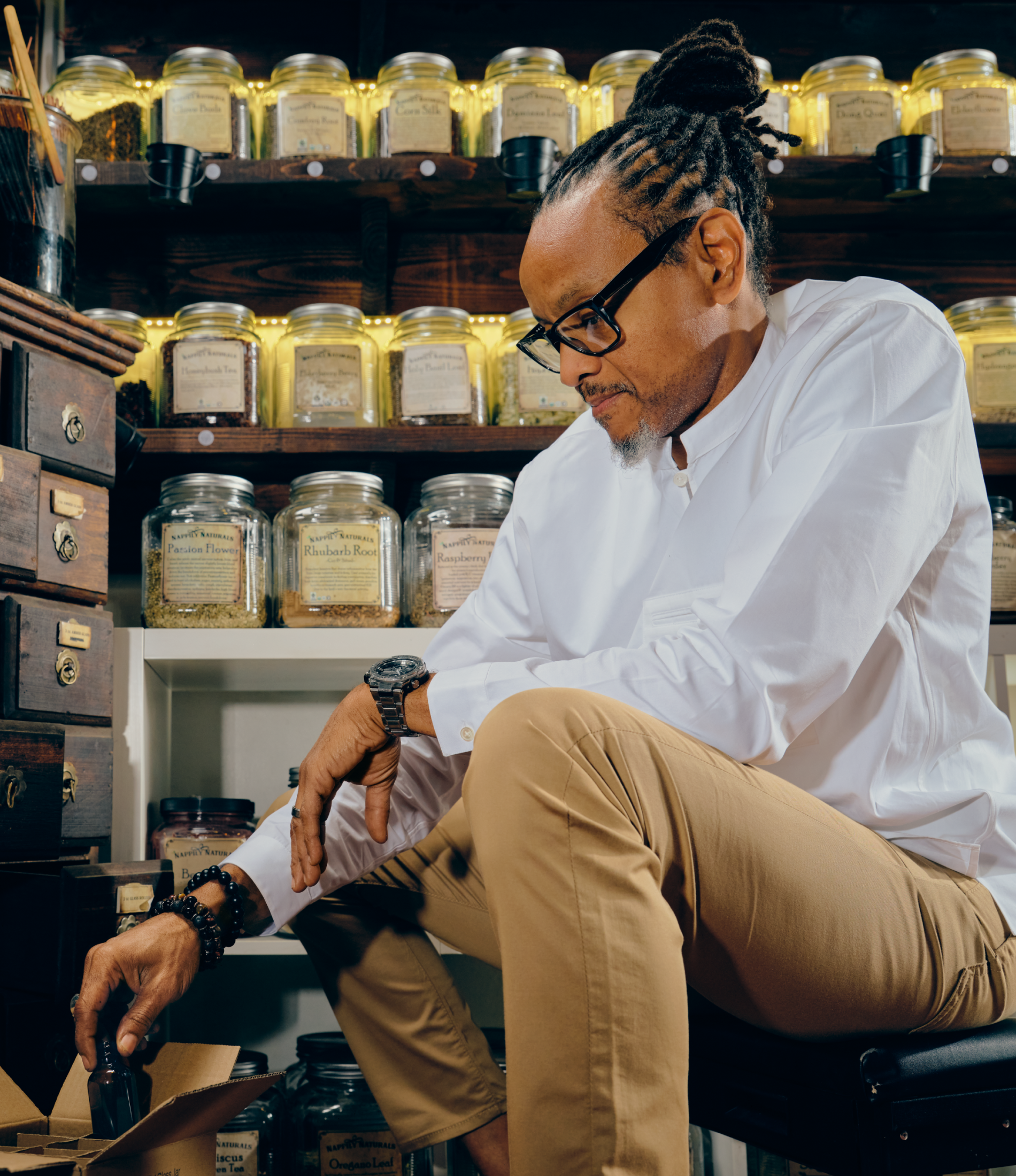 A person kneels on the ground looking at items for inventory in front of a wall with jars of loose-leaf herbs.