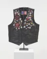 In a vertical image, a black leather vest is adorned with colorful pins across the front of it. Embroidery on the vest reads: "Felícitas "La Prieta" Méndez" 