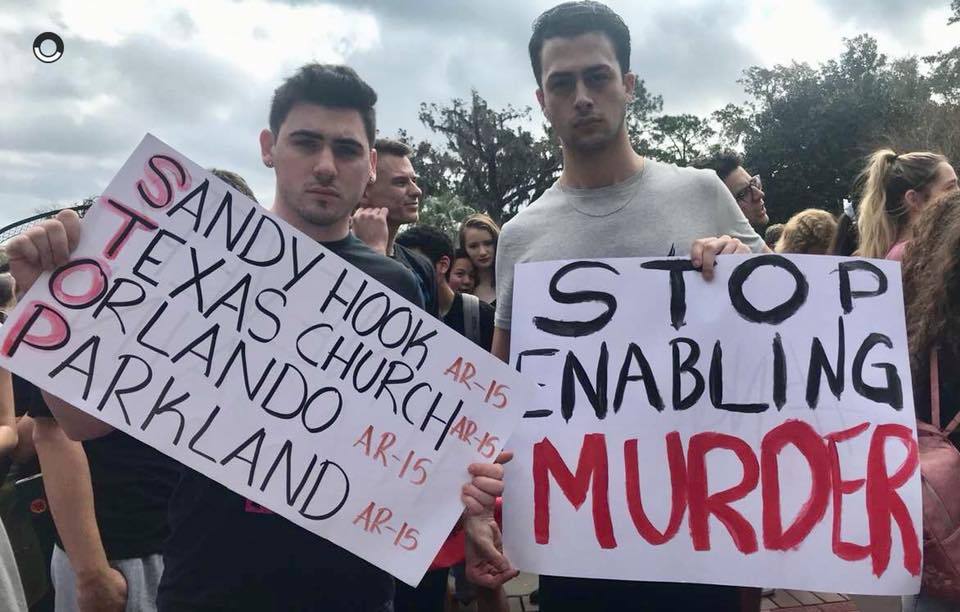 John (left) and Sean (right), two students of Florida State University, hold up homemade signs protesting gun violence. On the left, the sign adopts a diagnostic frame, acknowledging that the AR-15 was the designated weapon used for mass murders. The...