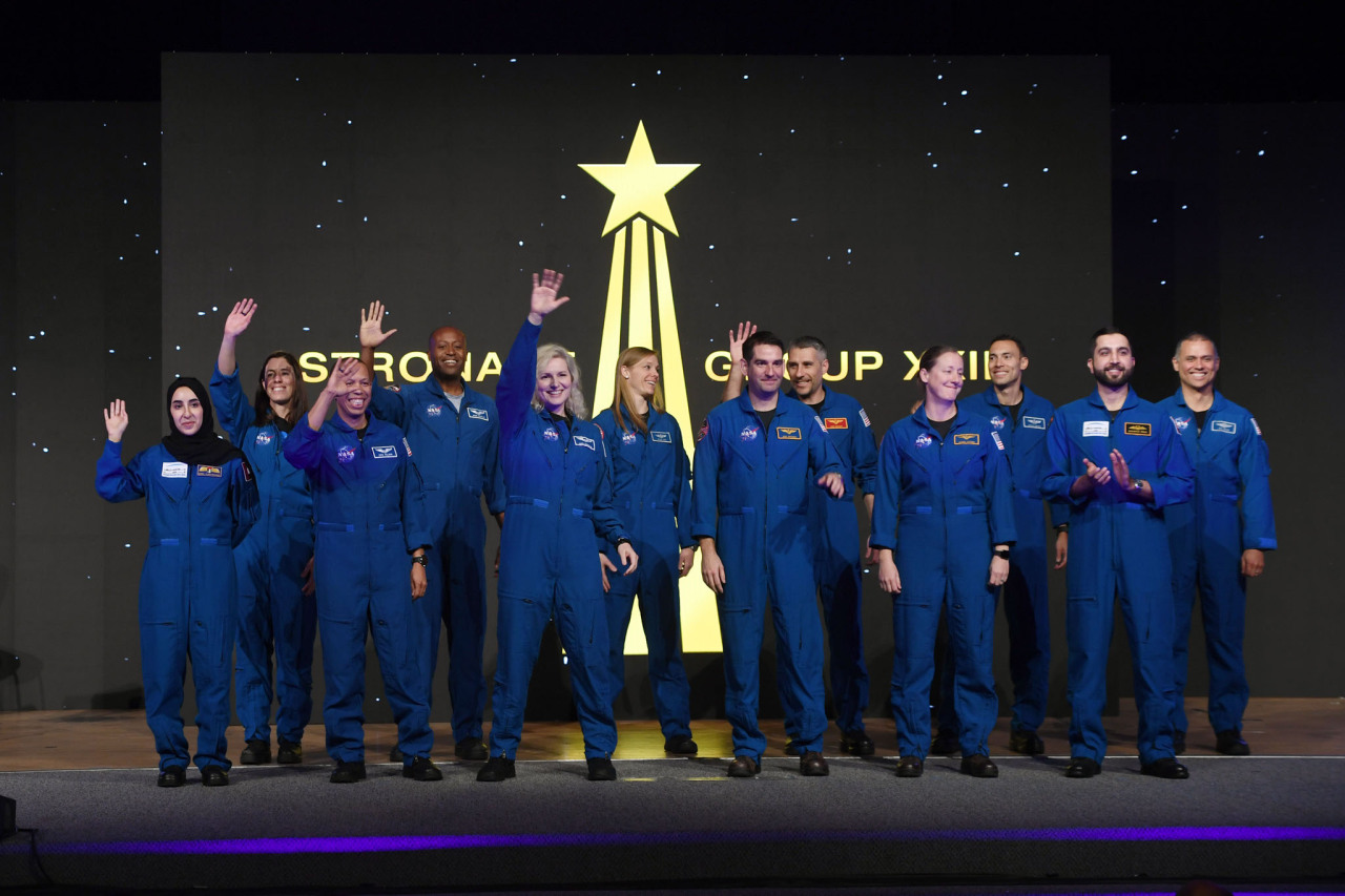 The latest astronaut candidate graduates, a group of men and women of different races and ethnicities, greet the audience (not pictured) at their graduation ceremony. The candidates all wear blue jumpsuits with patches on them. Behind them is a black and gold graphic of a star streaking upwards. The background has white dots on it that resemble distant stars. Credit: NASA