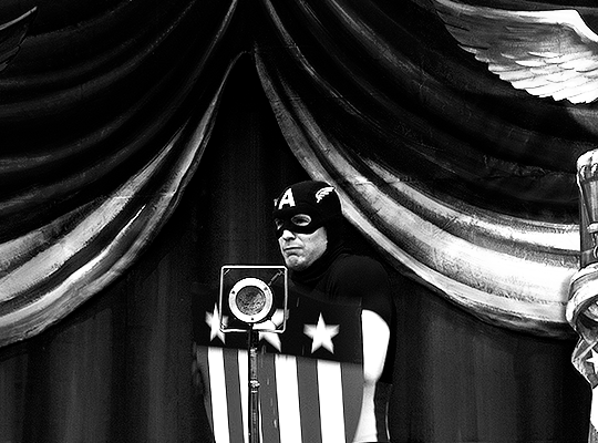 gif 8 of 9. a black and white gif. Steve dodges a tomato thrown at him. he's wearing his first Captain America outfit and he's on a stage. he's holding a small wooden shield, his first.