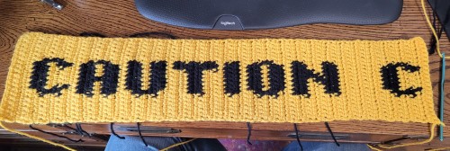 An in-progress crocheted scarf. It says "CAUTION C" in black text on a yellow background