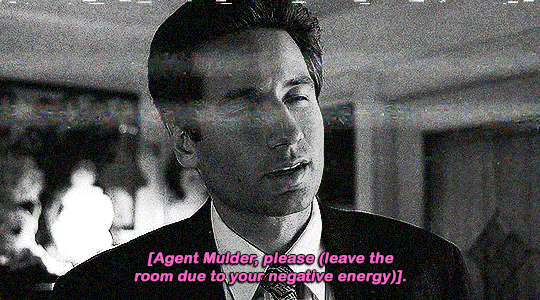 gif 2 of 14. Mulder has a sour expression after listening he has to leave the room due to his 'negative vibes'. Scully gets close to him and in a stage whisper, she says she can't bring him anywhere. the gif is in black and white and has a VHS glitch overlay effect.