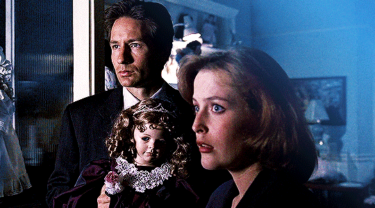 gif 5 of 14. Mulder and Scully both look at something intensively. Mulder is holding a vintage baby doll. the color blue is prominent in the gif.
