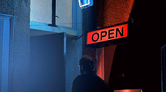 gif 9 of 14. a man goes inside a fortune teller shop. the camera pans to the shop's open sign and a neon sign of a hand with an eye in it. the gif's prominent colors are red and blue.