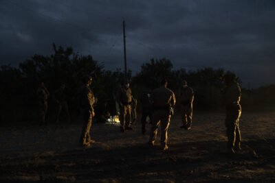 Five people stand in a loose circle on sandy ground. It's night and they are only faintly lit a light coming in from the right.