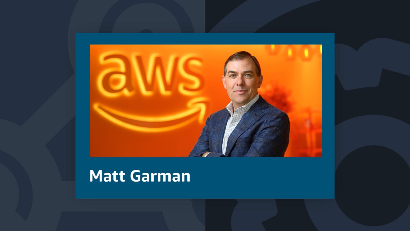 A photo of AWS CEO Matt Garman with his arms crossed. Garman is standing in front of a wall that has a lit sign of the AWS logo.
