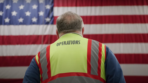 An image of an Amazon employee in a yellow work vest standing in front of an American flag.