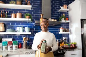 sylvia kapsandoy the founder of usimplyseason stands in front of a kitchen stove and countertop holding a carton of spice seasonings 