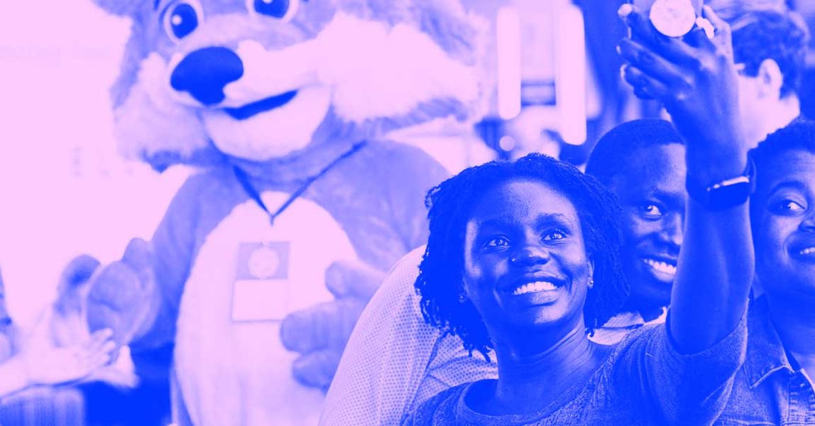 Three women stand in front of the Firefox mascot, Foxy, and take a selfie with him while other MozFest attendees cross in the background.