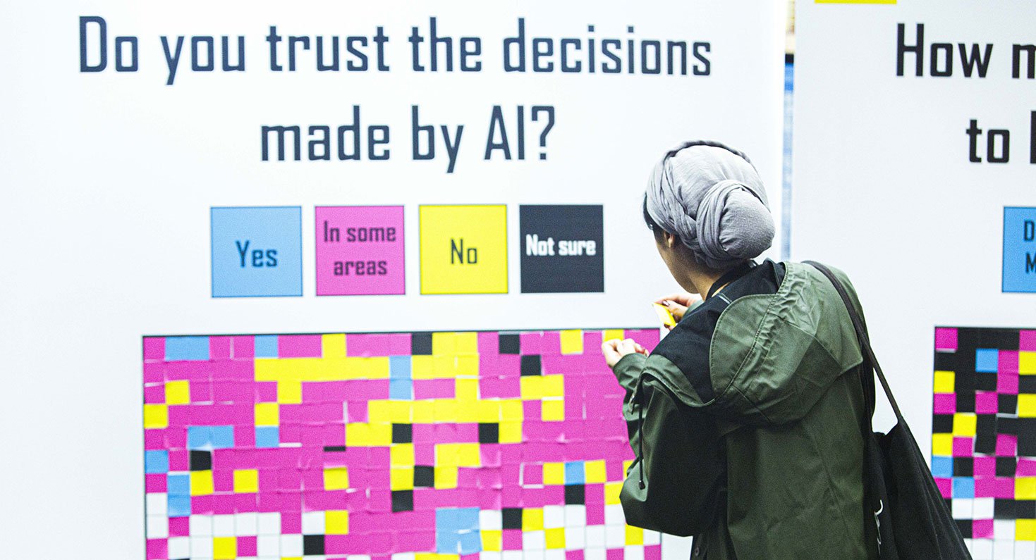 Een persoon voor een groot paneel vol notitieblaadjes, waarop staat ‘do you trust the decisions made by AI?’