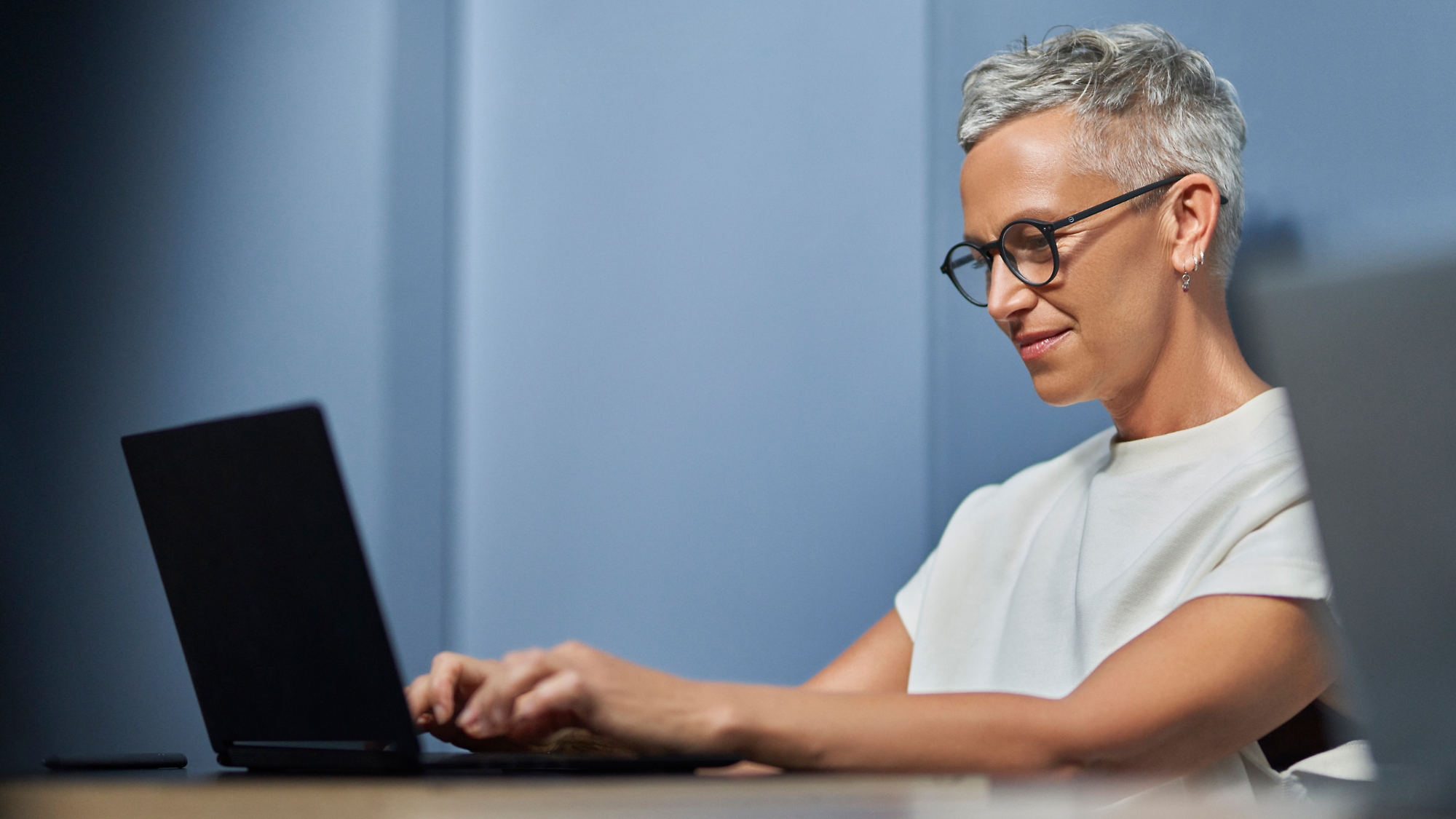 En moden kvinde med kort, gråt hår smiler og arbejder på en bærbar computer i et moderne kontormiljø.