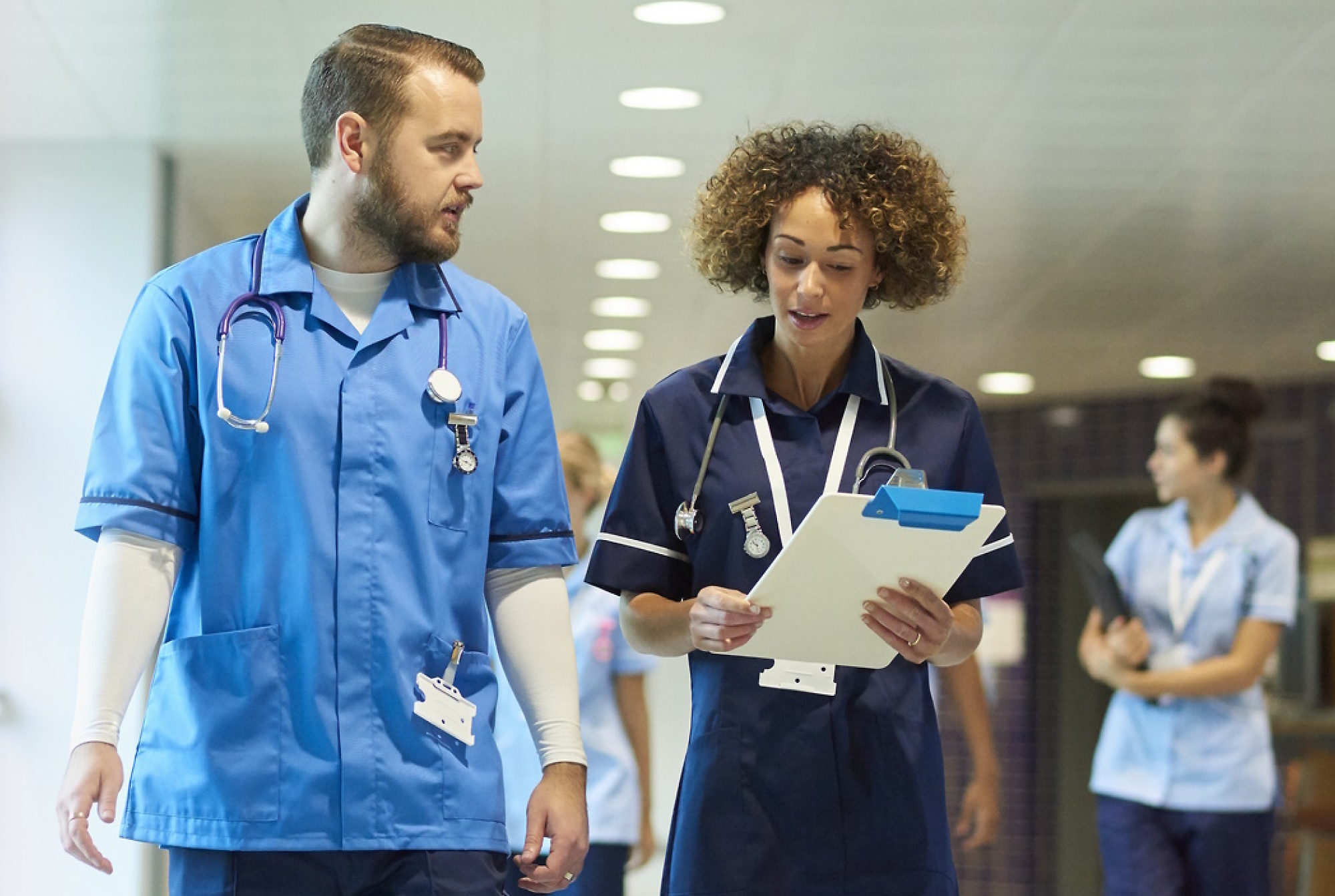 Two healthcare professionals in blue uniforms talking and walking in a hospital corridor, with one holding a clipboard. 