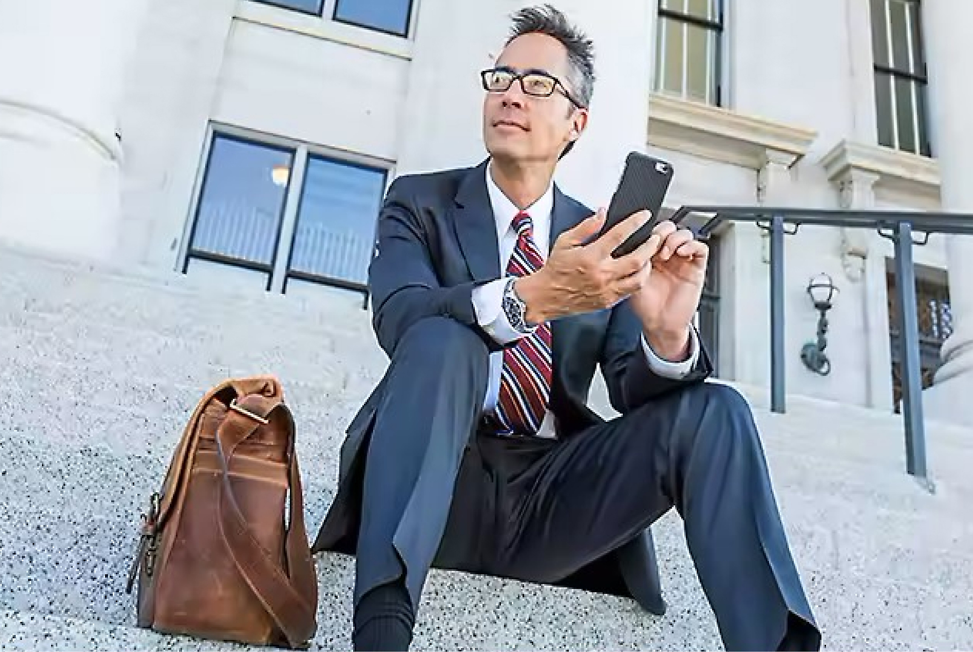 Podnikatel v obleku a brýlích sedí na schodech před budovou, používá smartphone a vedle sebe má koženou tašku.