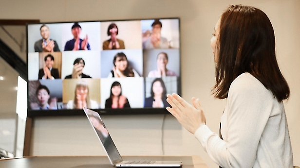 Mulher se apresentando a colegas por meio de uma chamada de videoconferência exibida em uma tela grande em um escritório moderno.