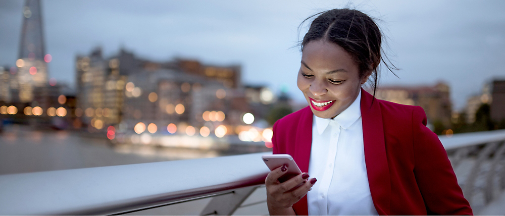 En smilende ung kvinde iført en rød blazer, der bruger sin smartphone på en bybro ved tusmørke.
