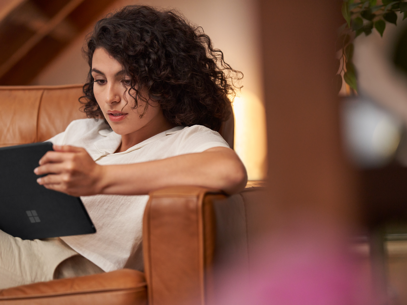 אישה מתולתלת קוראת בקשב רב בטאבלט, ישובה על כורסת עור חום בבית.