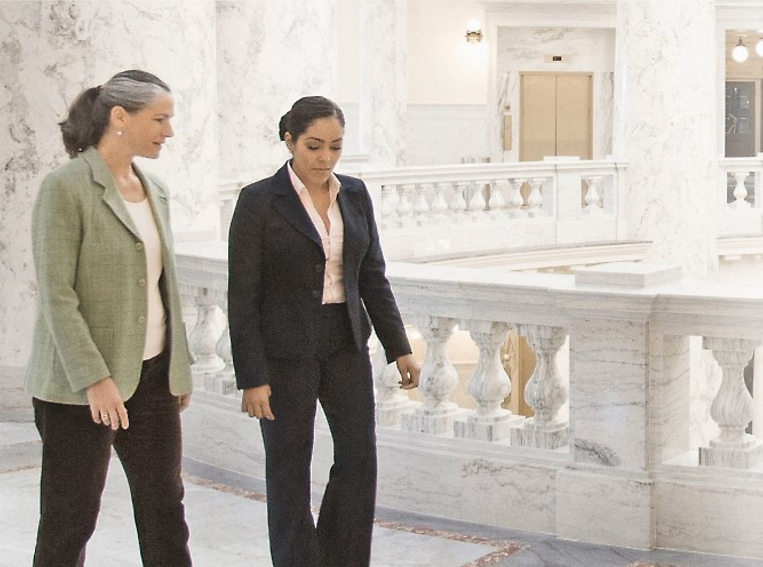 Duas mulheres caminhando e conversando em um salão de mármore com balaustradas ornamentadas em segundo plano.