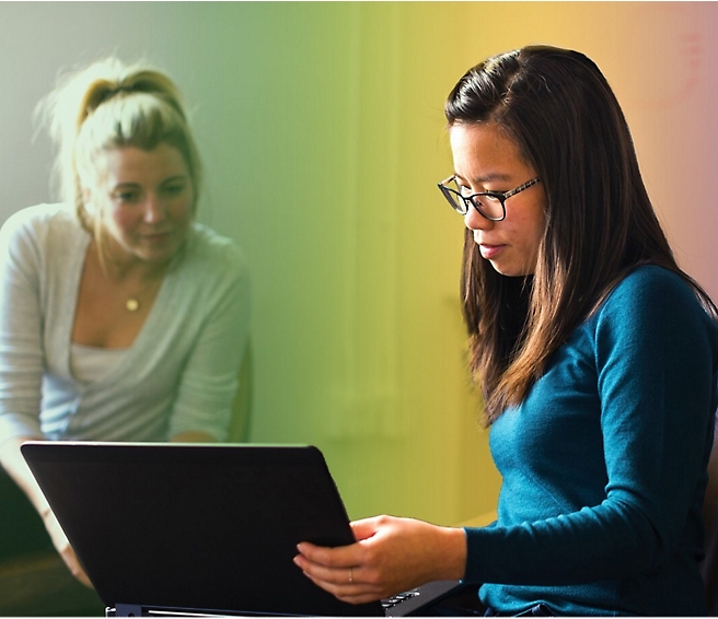 Две женщины в непринужденной офисной обстановке; одна работает на ноутбуке, а другая внимательно смотрит