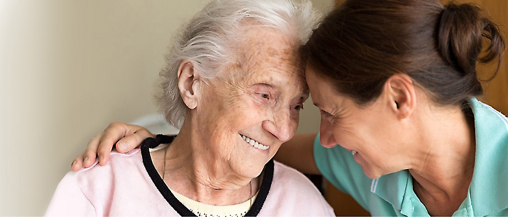En omsorgsperson som smiler til en eldre kvinne som også smiler, i varme innendørs omgivelser.