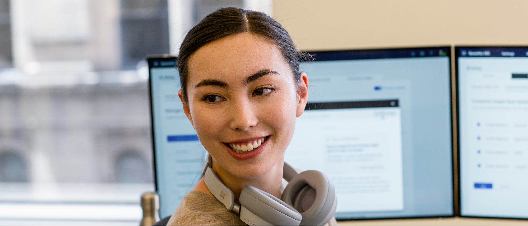 Een vrouw heeft haar koptelefoon om haar nek en werkt glimlachend op een paar monitoren.