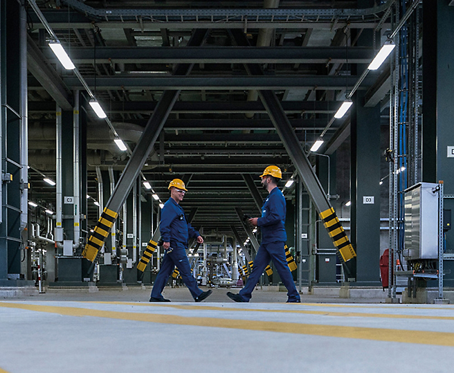 Δύο εργαζόμενοι με κράνη που περπατούν σε μια βιομηχανική εγκατάσταση.