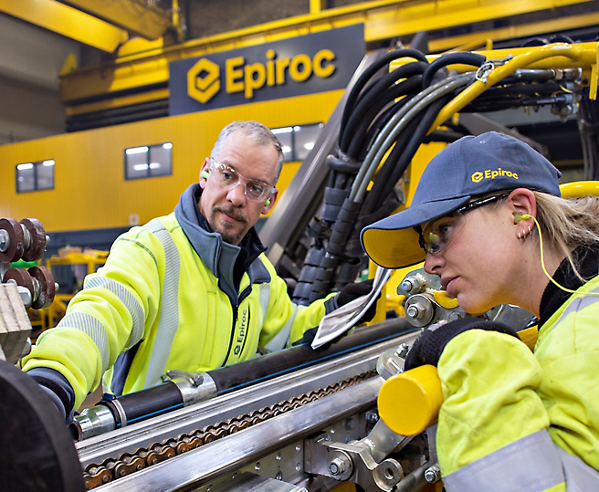 Epiroc 시설의 산업용 기계에서 작업하는 높은 가시성의 자켓을 입은 두 기술자.
