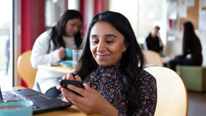 Personne souriante envoyant un message texte tout en utilisant un ordinateur portable dans une cafétéria.