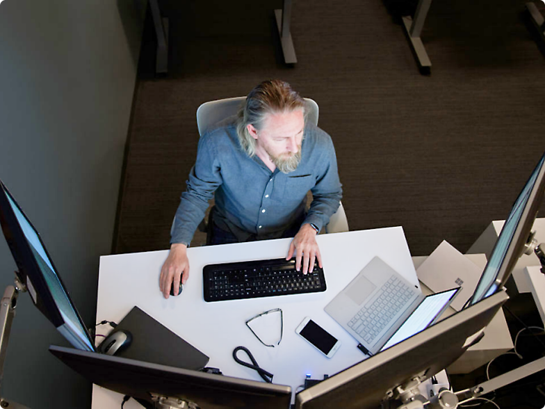 Ein Mann sitzt an einem Büroarbeitsplatz und benutzt eine Tastatur mit mehreren Monitoren und einem Laptop auf dem Schreibtisch.