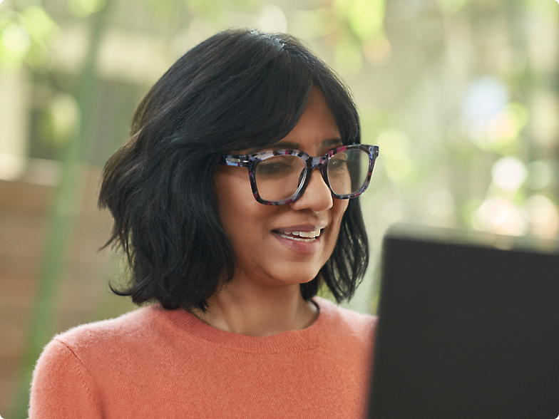 一名戴眼鏡穿著桃紅色毛衣的女士，在陽光照耀的房間裡，微笑地看著膝上型電腦螢幕。