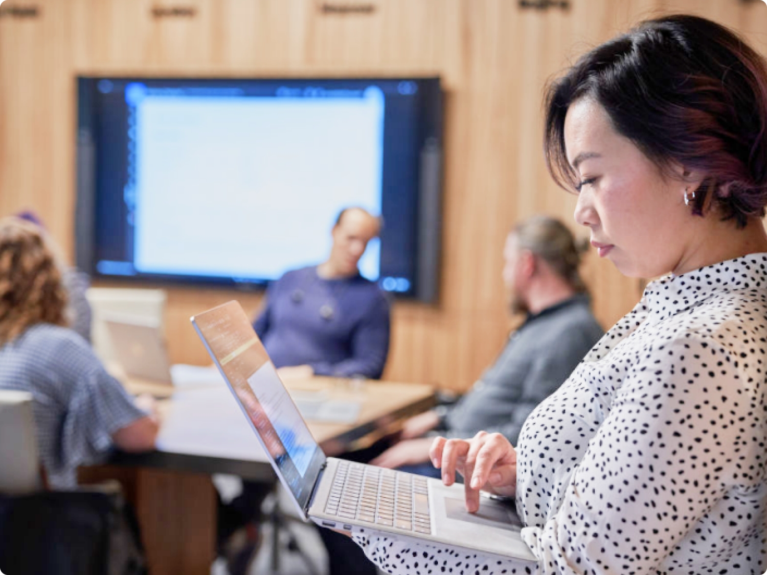一位女士在会议室中使用笔记本电脑，背景是其他三个同事和演示屏幕。