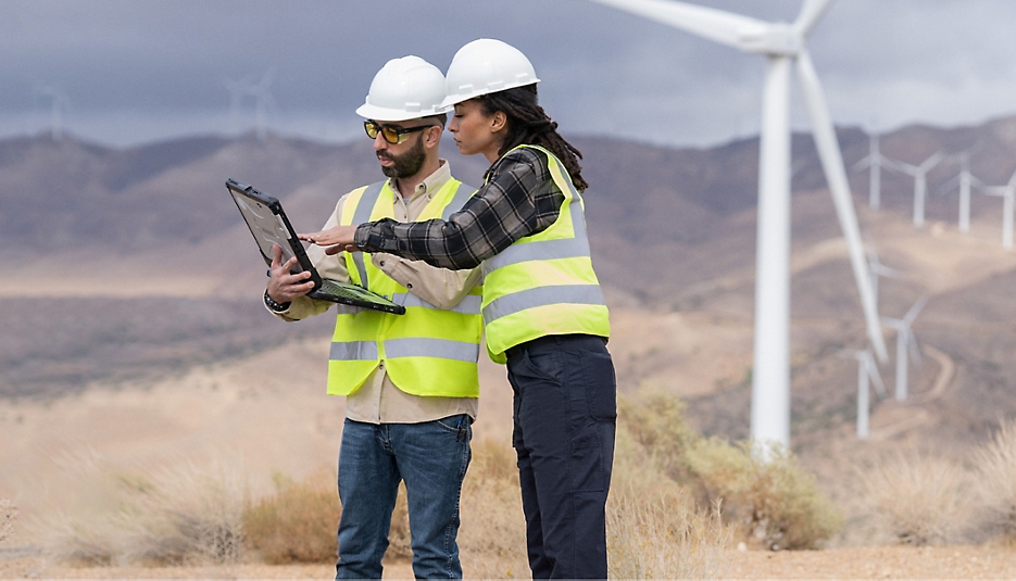 Two people wearing hard hats and safety vests standing in a field with wind turbines looking at a laptop