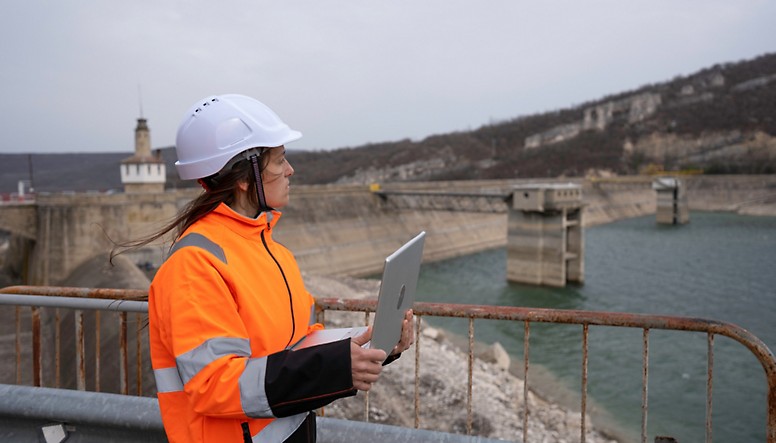 Um trabalhador da construção civil segura um tablet em frente a uma barragem.