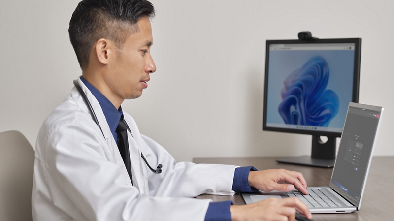 Una persona que trabaja en el ámbito sanitario lleva una bata blanca y un fonendoscopio, está sentada ante un escritorio y usa un portátil conectado a un monitor de escritorio