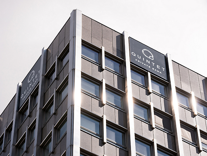 맑은 하늘 아래의 창문에 햇빛이 반사되는 "quintet" 로고가 있는 회색의 현대식 사무실 건물.