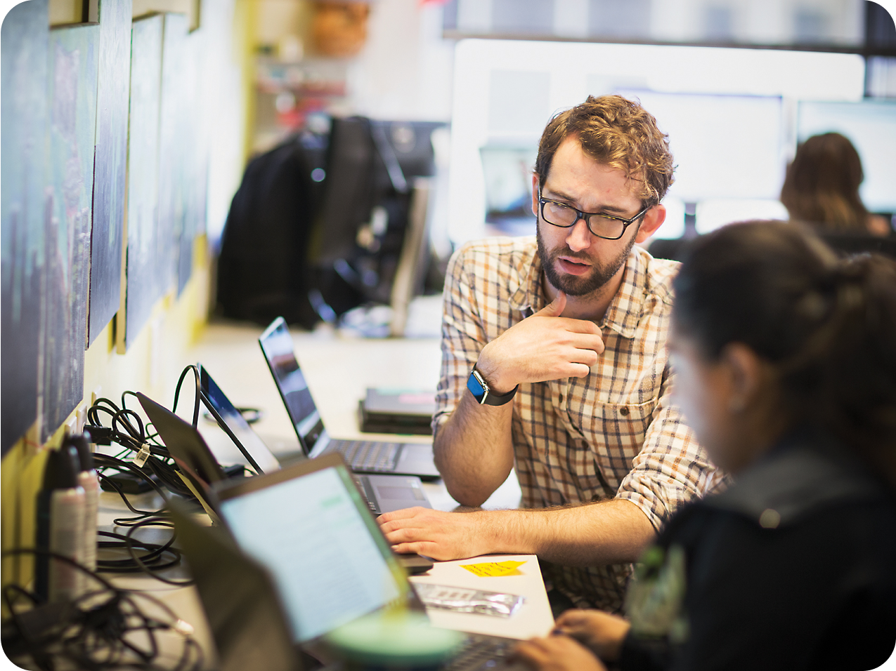 一位戴眼镜、留着胡须的男士在与一位女同事讨论时做出手势，两人都在繁忙的办公环境中使用笔记本电脑。