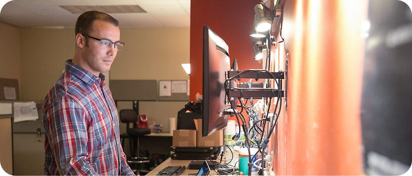 一位戴着眼镜、穿着格子衬衫的男士在光线昏暗的办公环境中在多台显示器上工作。