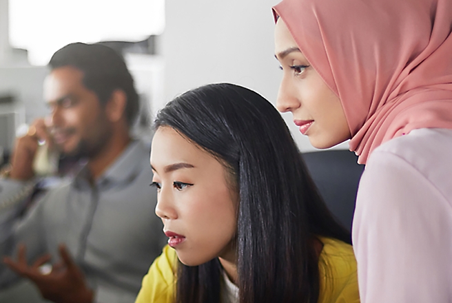 Two women are focused on a computer screen, with one woman wearing a hijab and the other looking intently at the screen.
