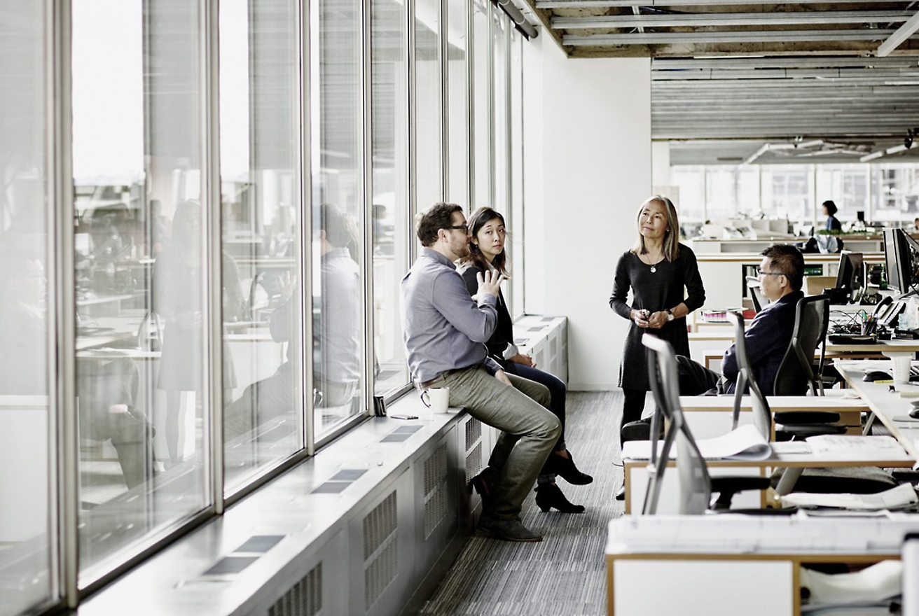 Four people in business attire have a discussion in a modern office, with three sitting on a ledge by large windows and one standing.