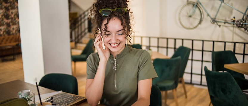A smiling woman with curly hair talking on her phone while sitting at a café table. 