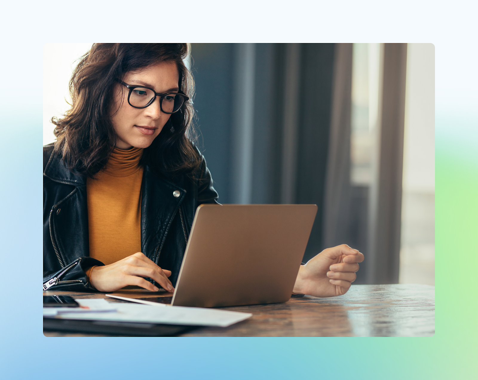 Eine Frau mit Brille, die sich auf die Arbeit an einem Laptop konzentriert, und an einem Schreibtisch mit Papieren sitzt, und eine schwarze Jacke trägt.