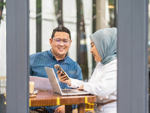 Zwei Personen sitzen in einem Café, sie lächeln und arbeiten an einem Projekt. Eine Person in einem blauen Hemd hält ein Dokument in der Hand, während die andere einen Hidschab trägt.