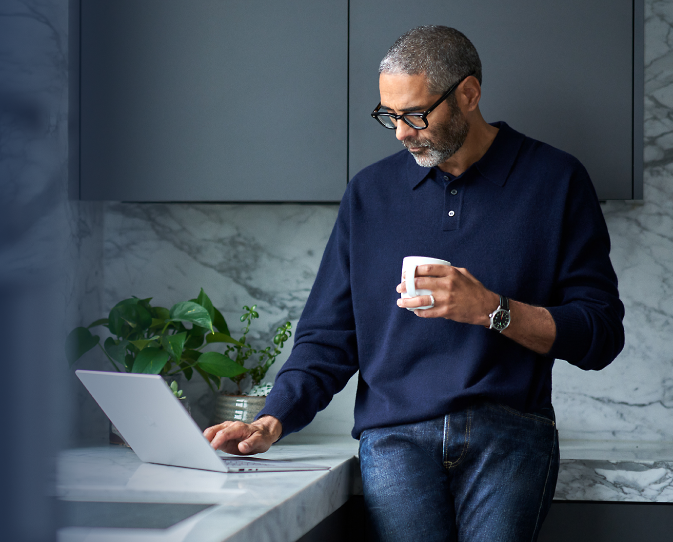 戴着眼镜的中年男子，手持咖啡杯，在现代厨房中使用笔记本电脑。