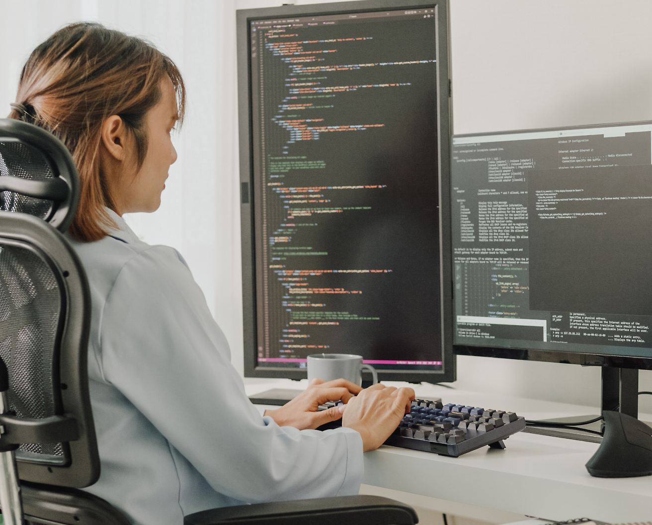 Bir ofis sandalyesinde oturmuş, programlama kodu gösteren birden fazla ekranla bilgisayarda kod yazan bir kadın.
