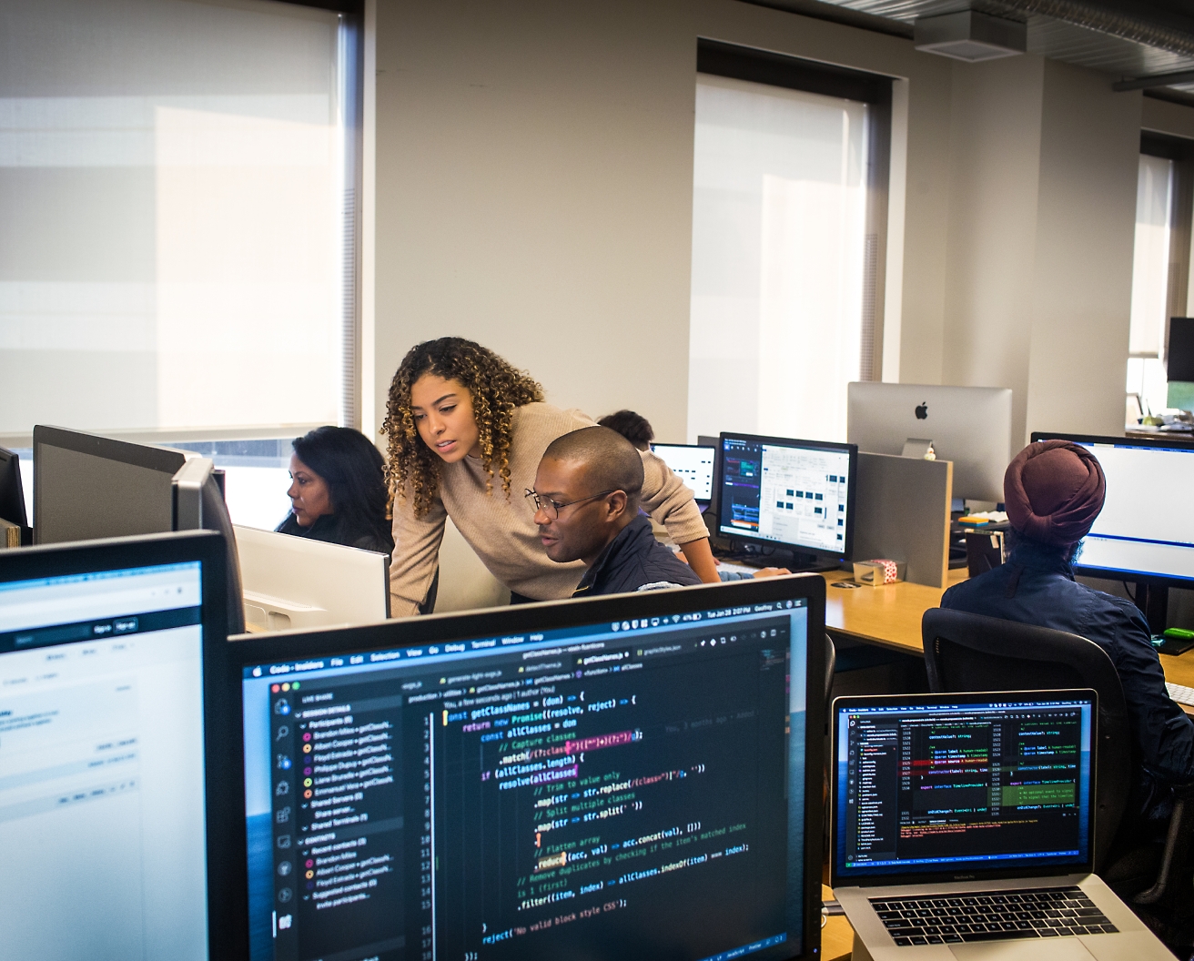 さまざまなコードやデータが表示された画面を中心に、オフィス環境でコンピューターで作業するさまざまなプロフェッショナルたち。