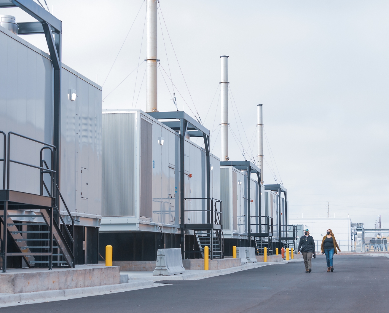 Bulutlu bir gökyüzü altında, bir tesiste uzun bacaları olan bir dizi endüstriyel konteynerin yanında yürüyen iki kişi.