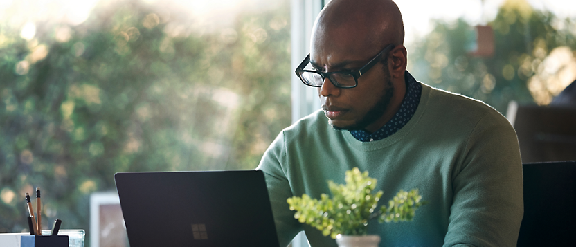 一位戴眼镜的男士专注地使用办公室上的笔记本电脑工作，背景是柔和的灯光，他旁边放着一盆小绿植。