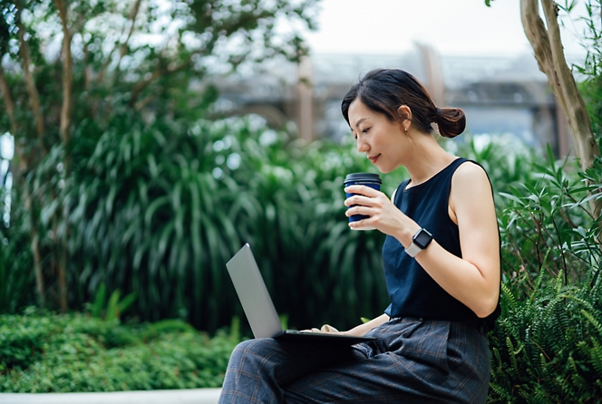 A woman sits outdoors on a bench with foliage behind her, holding a coffee cup in one hand and using a laptop