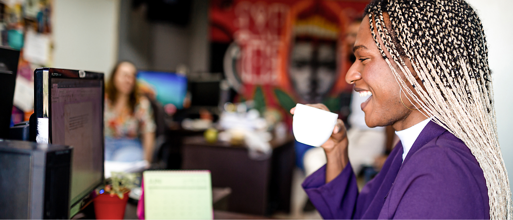 A person with braided hair, wearing a purple blazer, smiles while holding a cup in front of a computer in an office
