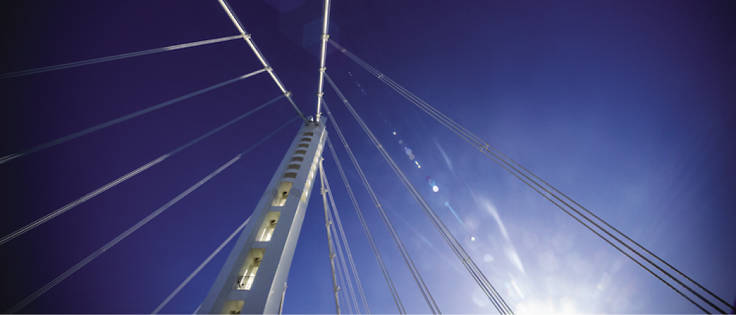 A close-up view of a modern cable-stayed bridge structure against a clear blue sky, with sunlight creating lens flares.